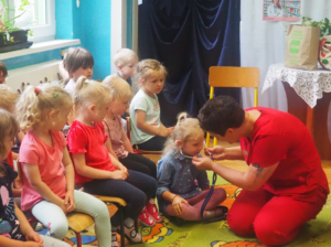 zdjęcie opisuje Panią pielęgniarkę z dziećmi, których uczy obsługi stetoskopu.