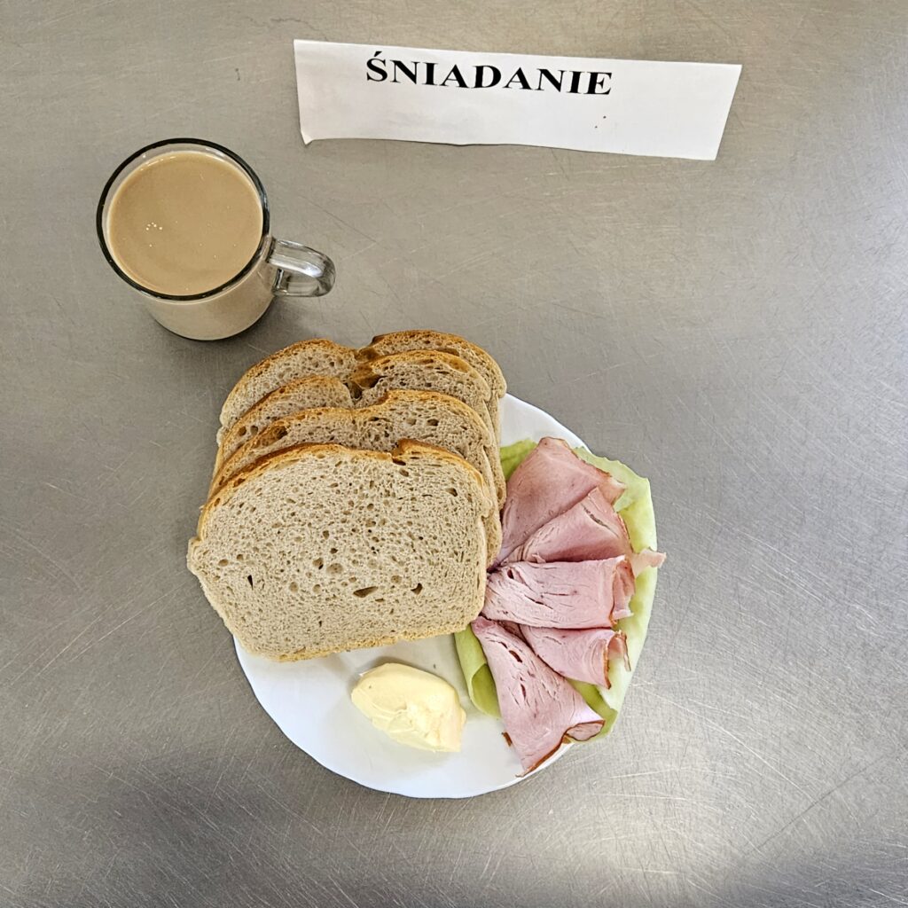 zdjęcie śniadania złożonego z: kiełbasy szynkowej, sałaty zielonej, chleba, margaryny oraz kawy z mlekiem.