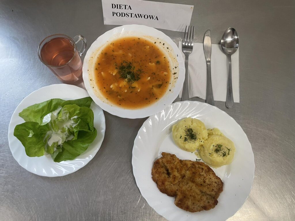 zdjęcie obiadu złożonego z: zupy pomidorowej z makaronem, kotleta drobiowego, ziemniaków, sałaty zielonej oraz kompotu.