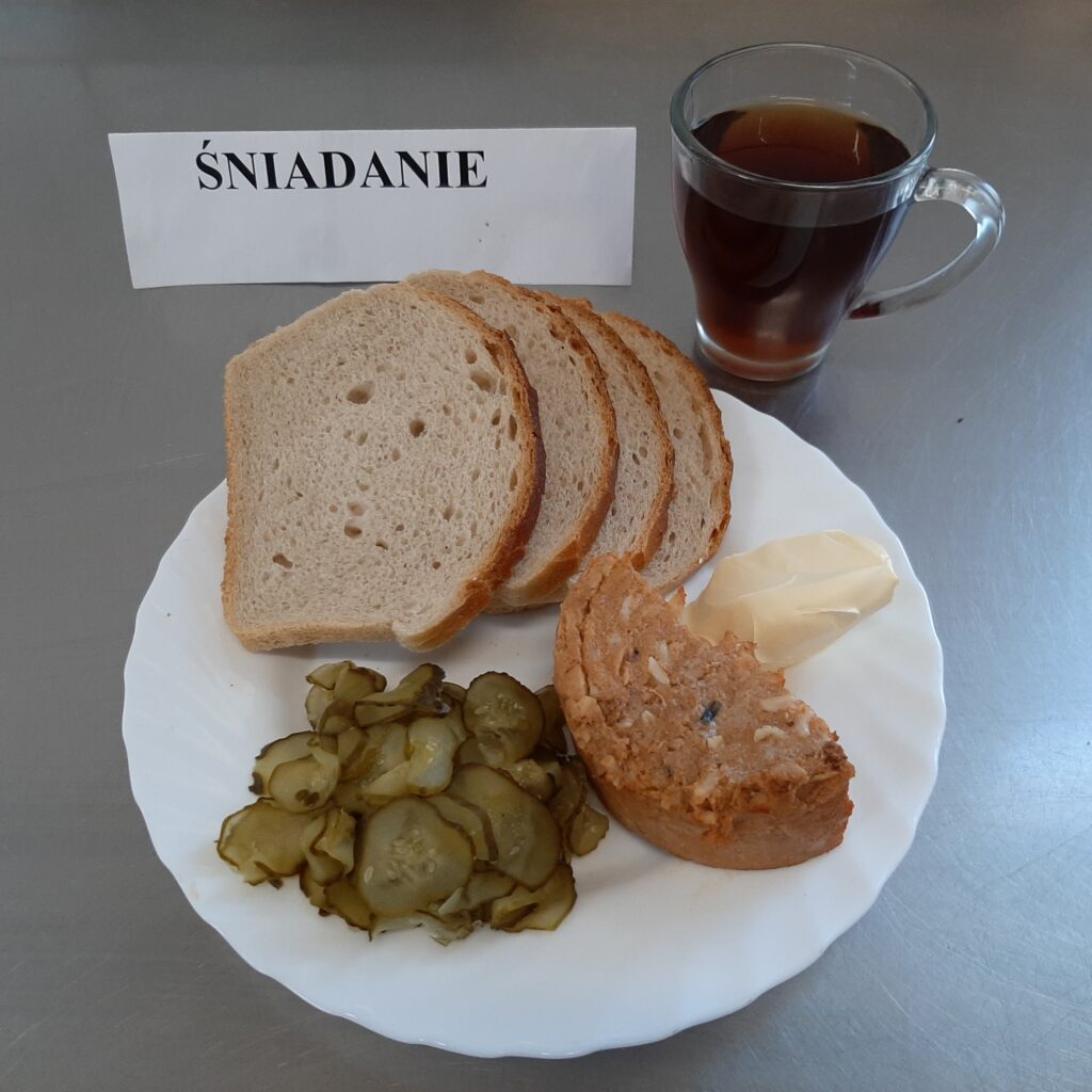 Zdjęcie śniadanie złożonego z: paprykarza szczecińskiego, ogórka kiszonego, chleba pszennego, margaryny oraz herbaty.