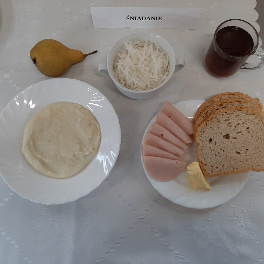 Zdjęcie śniadania złożonego z: chleba mazowieckiego, margaryny, szynki drobiowej, białej rzodkiewki, zupy mlecznej z kaszą manną, gruszką oraz herbatą.