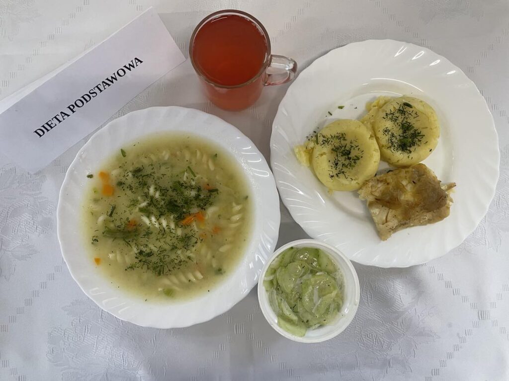 zdjęcie obiadu złożonego z: zupy jarzynowej z kluskami, ziemniakami puree, kompotu z jabłek, ryby pieczonej, piernika oraz mizerii.