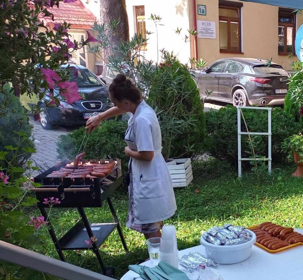 zdjęcie przedstawia pięknie przystrojony ogródek oraz grilla już z położonymi na niego kiełbaskami którymi zajmuje się Pracownica Naszego Szpitala.