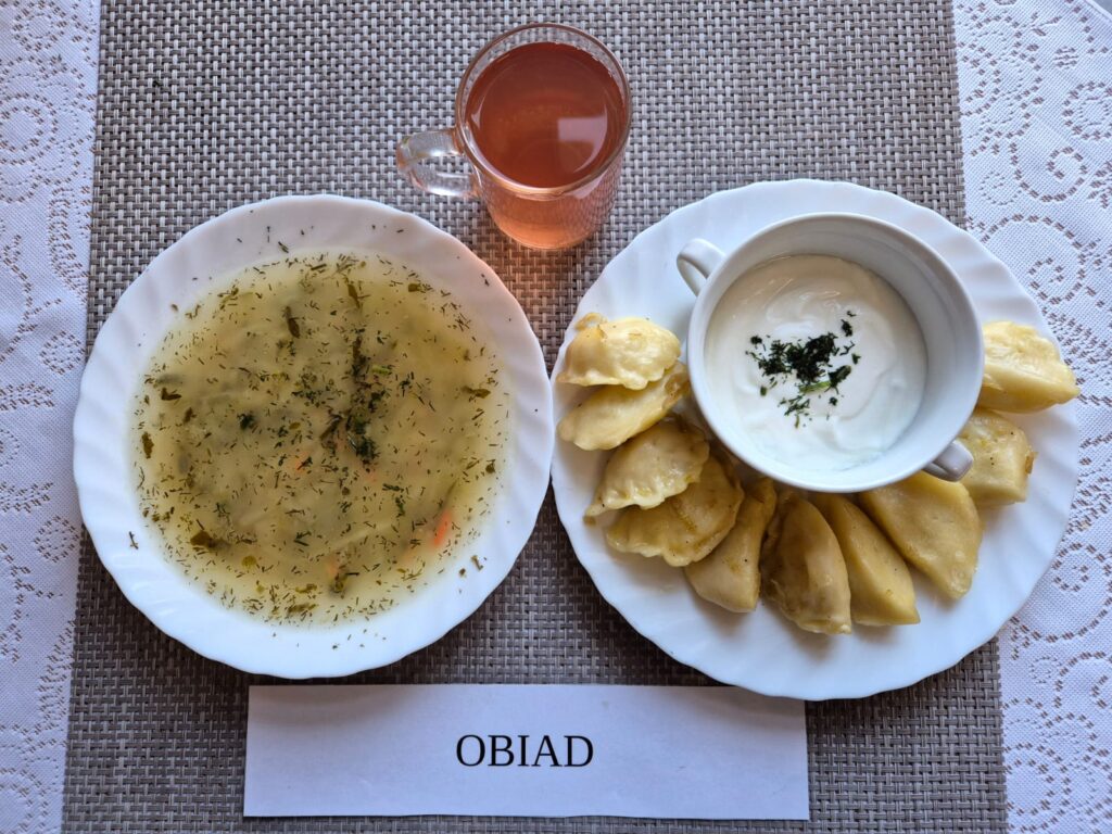 Na zdjęciu znajduję się posiłek w którego skład wchodzi Zupa ogórkowa z ziemniakami, Pierogi, kefir oraz kompot z jabłek