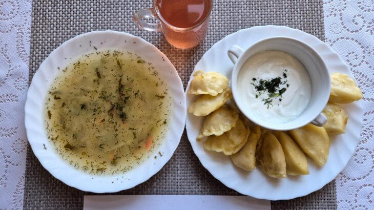 Na zdjęciu znajduję się posiłek w którego skład wchodzi Zupa ogórkowa z ziemniakami, Pierogi, kefir oraz kompot z jabłek