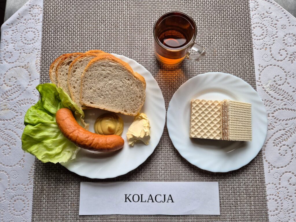 Na zdjęciu znajduję się posiłek w którego skład wchodzi, Chleb pszenny, Margaryna Śniadaniowa, Kiełbasa parówkowa, musztarda, wafle tortowe oraz Herbata czarna