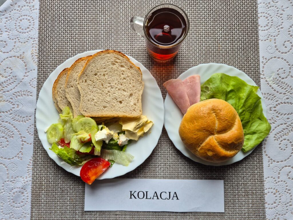 Na zdjęciu znajduję się posiłek w którego skład wchodzi Chleb pszenny, Margaryna Śniadaniowa, Sałatka grecka, Kajzerka, wędlina oraz Herbata czarna