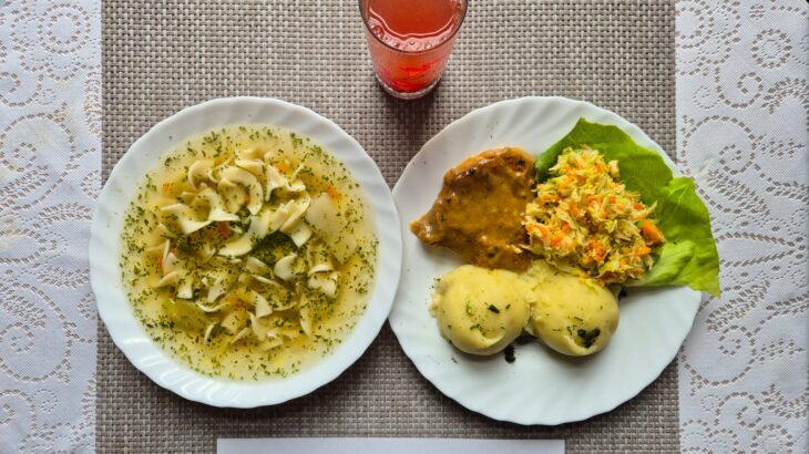 Na zdjęciu znajduję się posiłek w którego skład wchodzi Zupa makaronowa, Ziemniaki, Bitka wieprzowa, Surówka colesław oraz Kompot z jabłek