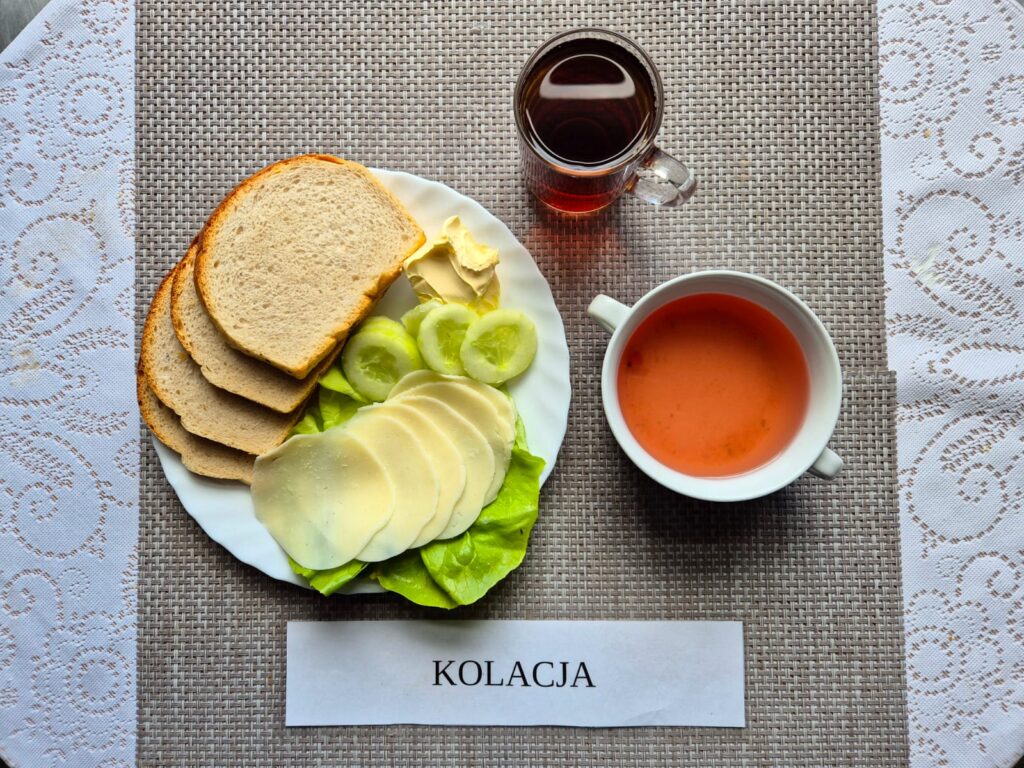 Na zdjęciu znajduję się posiłek w którego skład wchodzi Chleb pszenny, Margaryna śniadaniowa, Ser twardy, Sałata masłowa, ogórek zielony, kisiel oraz Herbata czarna