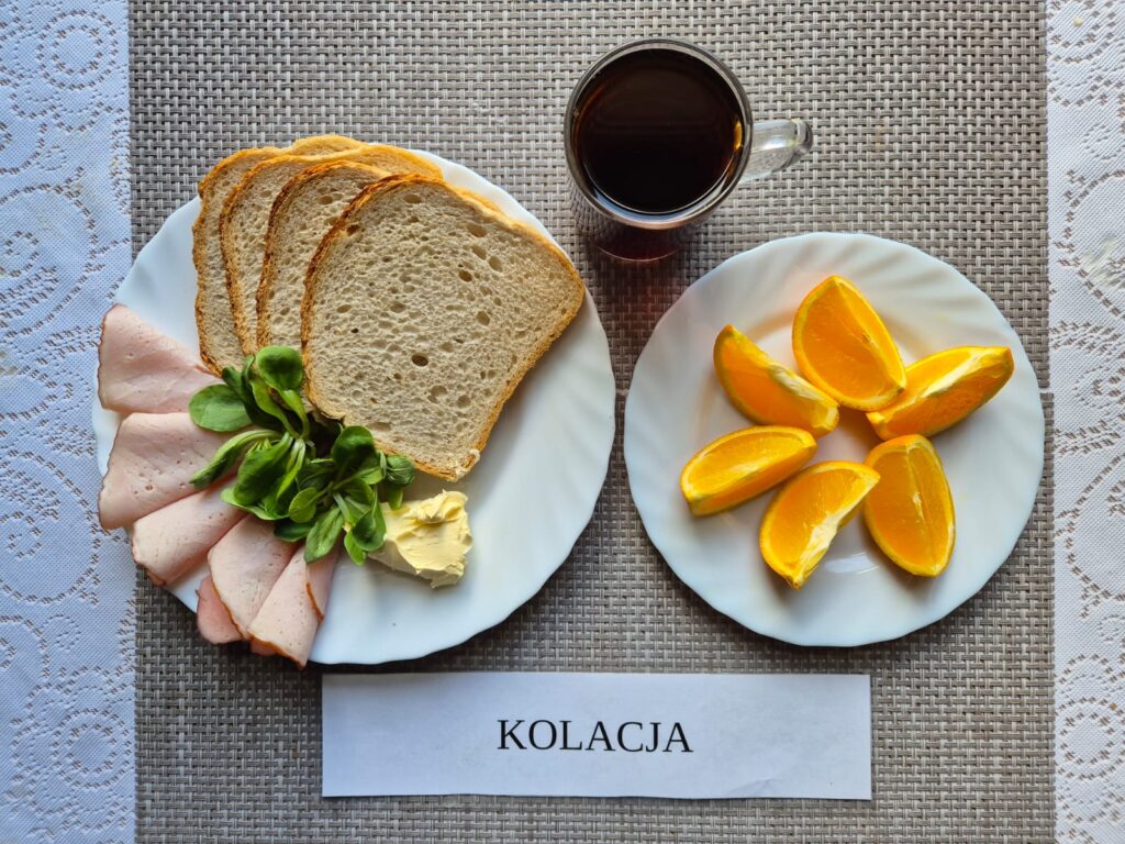 Na zdjęciu znajduję się posiłek w którego skład wchodzi Chleb pszenny, Margaryna Śniadaniowa, Kiełbasa krakowska, Sałata masłowa, pomarańcza oraz Herbata czarna