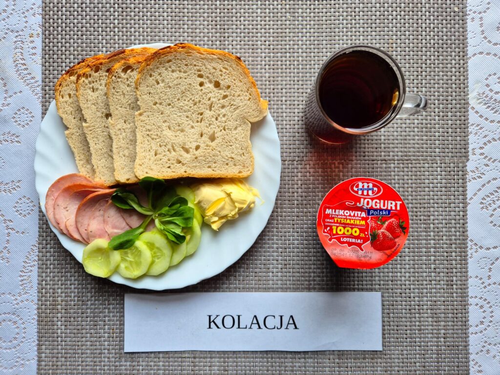 Na zdjęciu znajduję się posiłek w którego skład wchodzi Chleb pszenny, wędlina wieprzowa, ogórek świeży, Jogurt owocowy oraz herbata czarna
