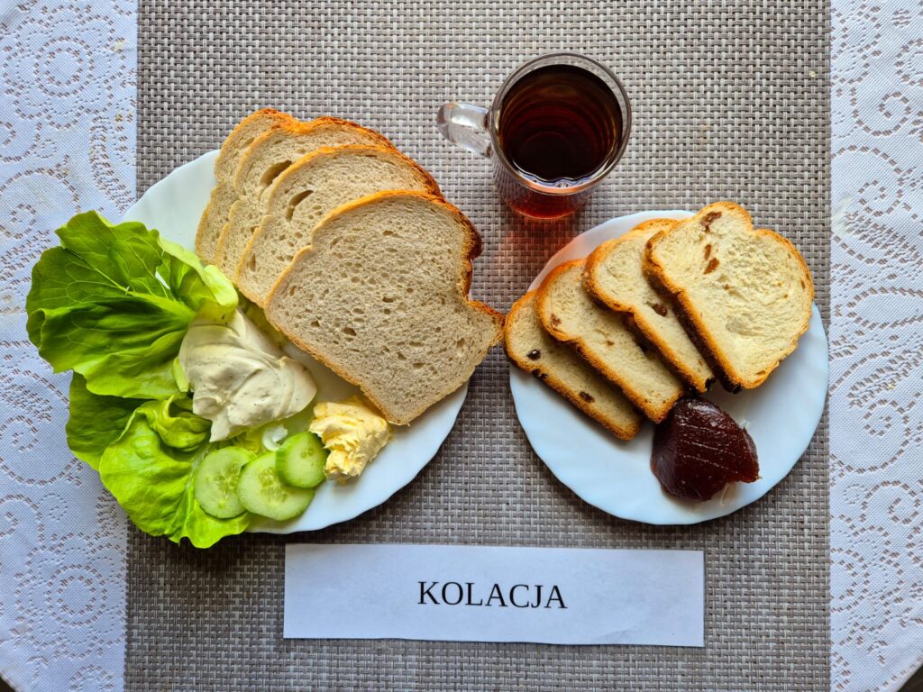 Na zdjęciu znajduję się posiłek w którego skład wchodzi Chleb pszenny, Margaryna Śniadaniowa, ser topiony, sałata masłowa, pomidor, Chałka, dżem oraz herbata czarna