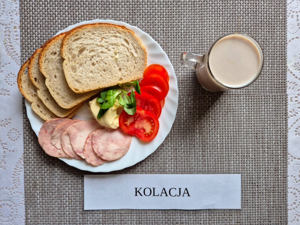 Na zdjęciu znajduję się posiłek w którego skład wchodzi Chleb pszenny, Margaryna Śniadaniowa, Kiełbasa krakowska drobiowa, pomidor oraz Herbata czarna