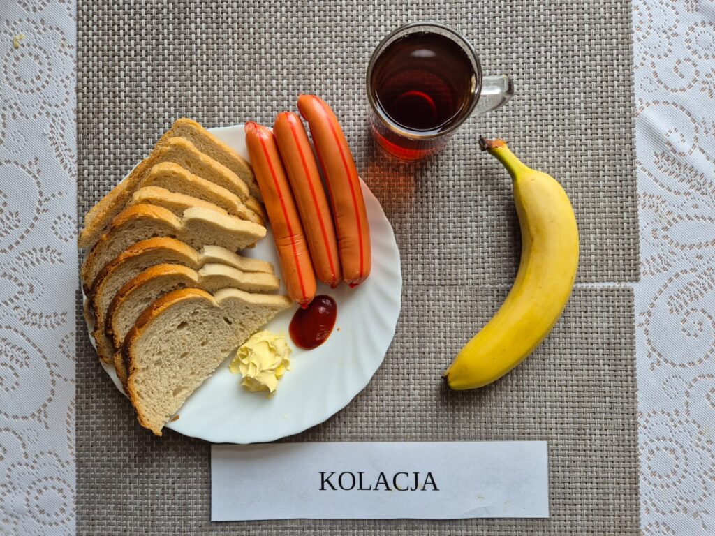 Na zdjęciu znajduję się posiłek w którego skład wchodzi Chleb pszenny, Margaryna Śniadaniowa, parówki drobiowe, Ketchup, banan oraz Herbata czarna