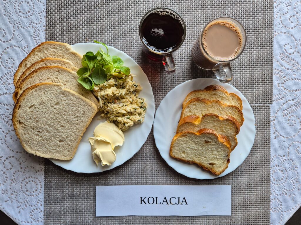 Na zdjęciu znajduję się posiłek w którego skład wchodzi Chleb pszenny, Margaryna Śniadaniowa, Pasta z jajka, ogórka konserwowego i pieczarek marynowanych z dodatkiem szczypiorku, kakao, chałka oraz Herbata czarna
