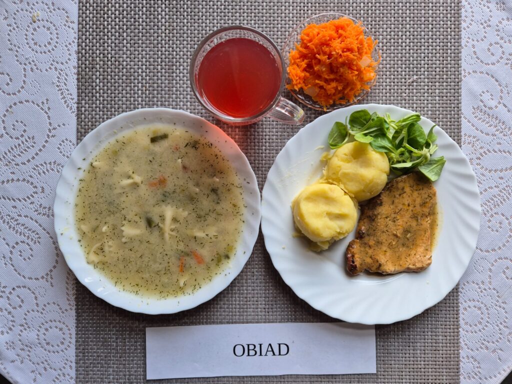 Na zdjęciu znajduję się posiłek w którego skład wchodzi Zupa porowa z makaronem, Ziemniaki, Bitka wieprzowa w sosie koperkowym, Surówka z marchewki z dodatkiem ananasa oraz Kompot z jabłek