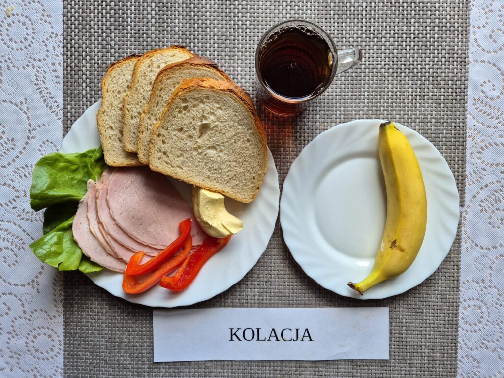 Na zdjęciu znajduję się posiłek w którego skład wchodzi Chleb pszenny, Margaryna Śniadaniowa, Kiełbasa krakowska drobiowa, sałata masłowa, papryka, banan oraz Herbata czarna