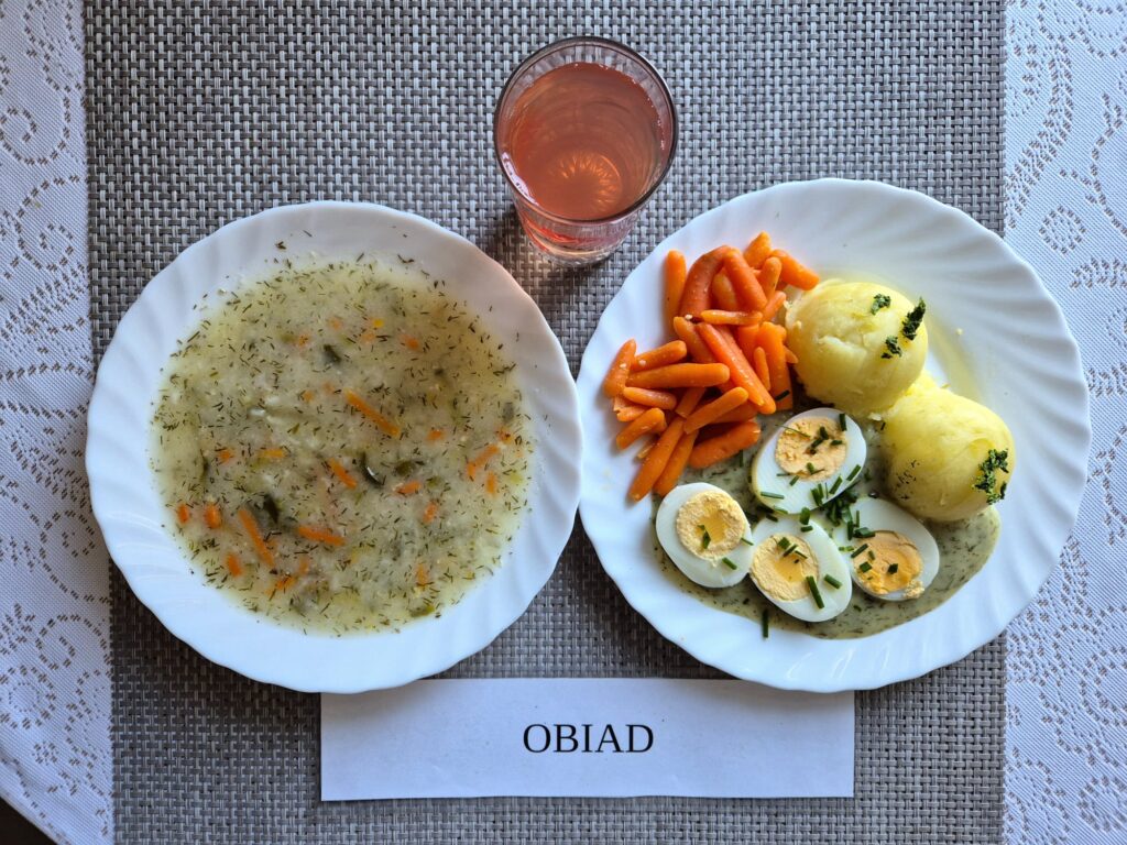 Na zdjęciu znajduję się posiłek w którego skład wchodzi Zupa ogórkowa z ryżem, Ziemniaki, Jajka 2 szt w sosie koperkowym, Marchewka mini oraz kompot z jabłek
