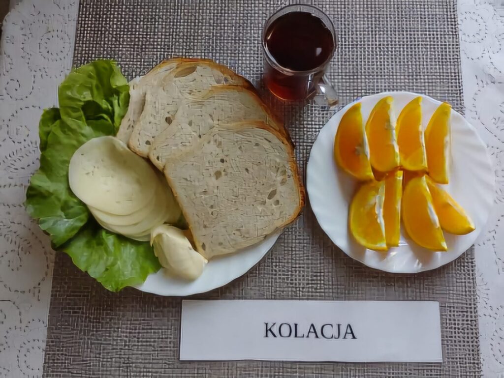 Na zdjęciu znajduję się posiłek w którego skład wchodzi Chleb pszenny, Margaryna Śniadaniowa, Ser twardy, sałata masłowa, pomarańcza oraz Herbata czarna