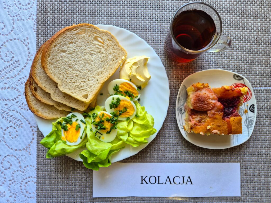 Na zdjęciu znajduję się posiłek w którego skład wchodzi Chleb pszenny, margaryna śniadaniowa, Jajka 2 szt. sosie majonezowym ze szczypiorkiem, Sałata masłowa, Ciasto z truskawkami oraz Herbata czarna