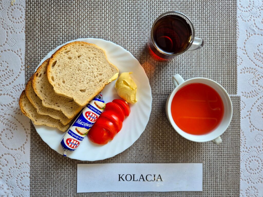 Na zdjęciu znajduję się posiłek w którego skład wchodzi Chleb pszenny, Margaryna Śniadaniowa, ser topiony, pomidor, kisiel oraz Herbata czarna