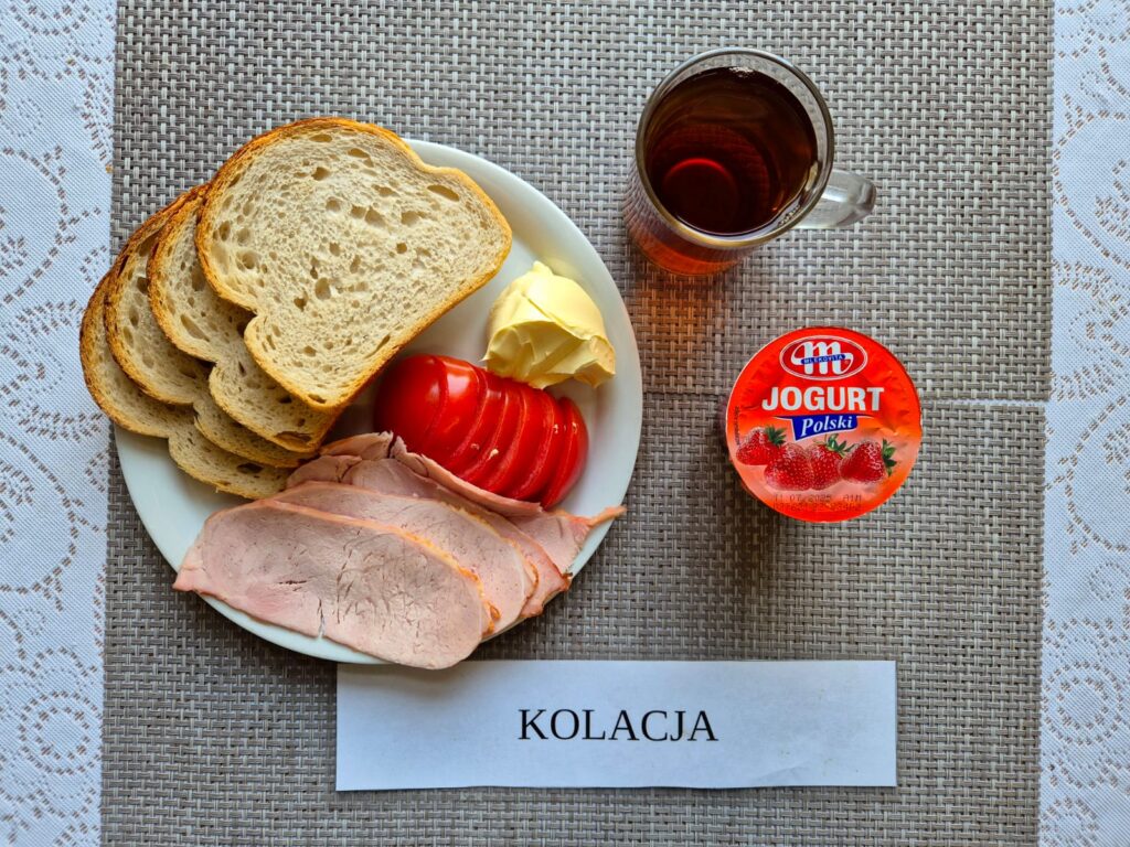 Na zdjęciu znajduję się posiłek w którego skład wchodzi Chleb pszenny, Margaryna Śniadaniowa, Wędlina wieprzowa, pomidor, jogurt owocowy oraz Herbata czarna