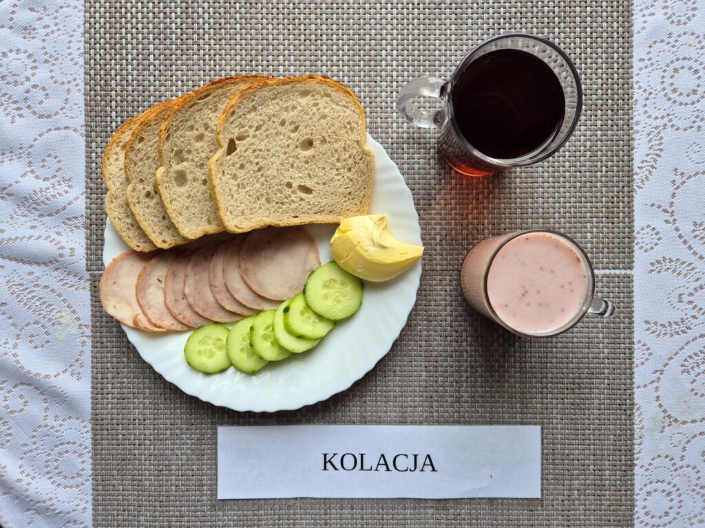 Na zdjęciu znajduję się posiłek w którego skład wchodzi Chleb pszenny, Margaryna Śniadaniowa, Kiełbasa krakowska drobiowa, ogórek zielony, koktajl truskawkowy oraz Herbata czarna
