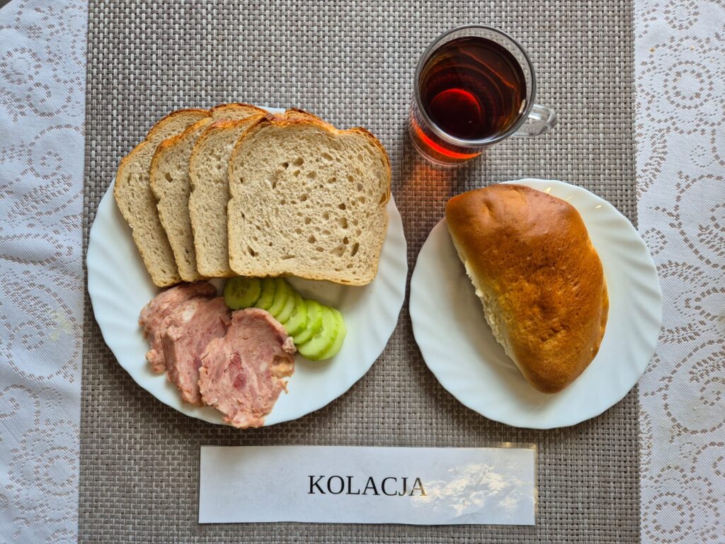 Na zdjęciu znajduję się posiłek w którego skład wchodzi Chleb pszenny, Margaryna Śniadaniowa, Ser topiony, Ogórek zielony, Drożdżówka oraz Herbata czarna