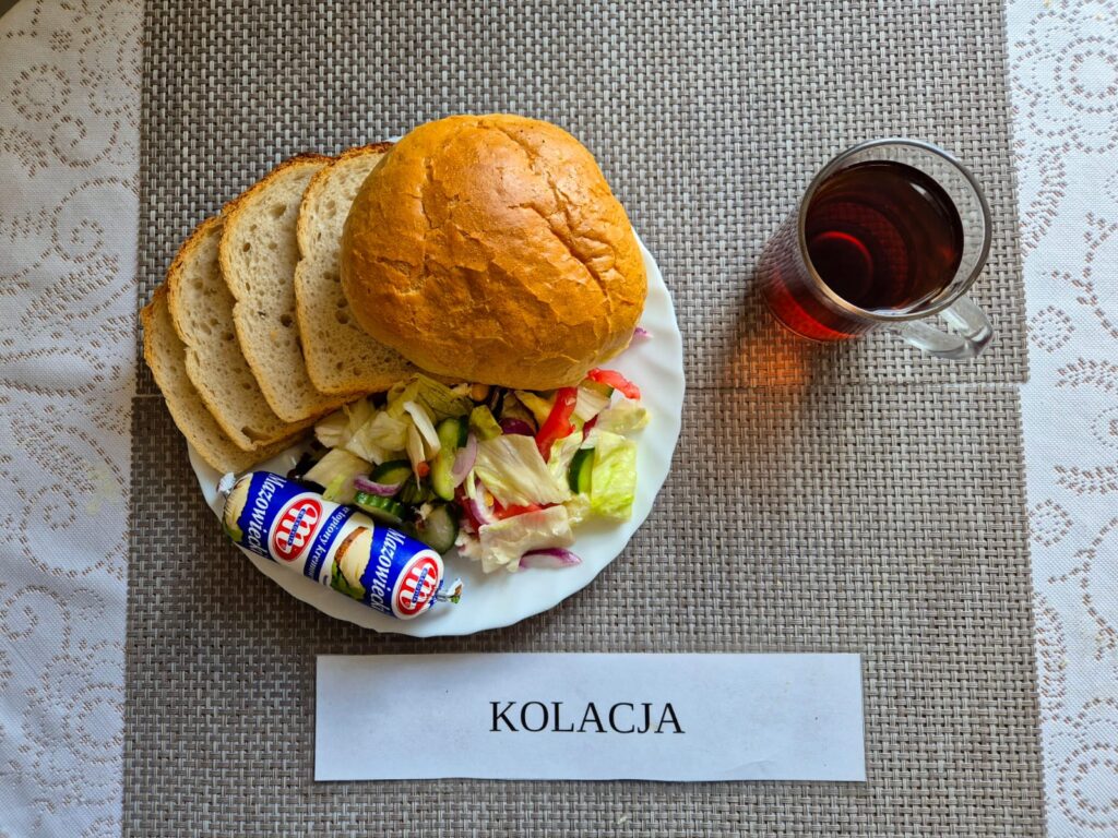 Na zdjęciu znajduję się posiłek w którego skład wchodzi Chleb pszenny, margaryna śniadaniowa, Sałatka grecka, ser topiony, bułka pszenna oraz Herbata czarna