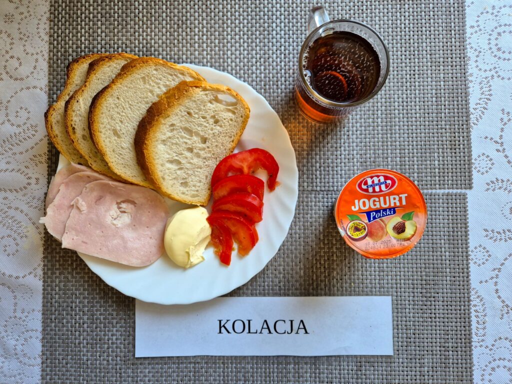 Na zdjęciu znajduję się posiłek w którego skład wchodzi Chleb pszenny, Margaryna Śniadaniowa, Wędlina wieprzowa, pomidor, jogurt owocowy oraz Herbata czarna
