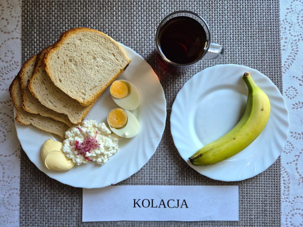 Na zdjęciu znajduję się posiłek w którego skład wchodzi Chleb pszenny, Margaryna Śniadaniowa, Twarożek z rzodkiewką i szczypiorkiem, jajko, pomidor, banan oraz Herbata czarna
