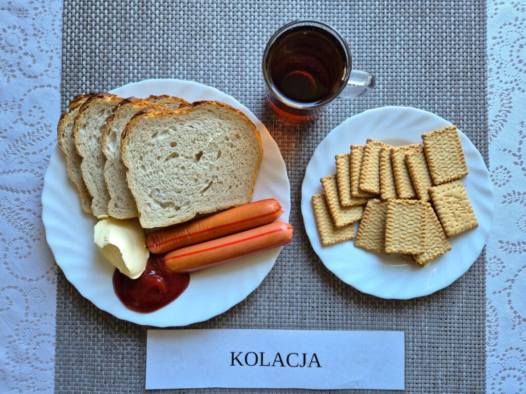 Na zdjęciu znajduję się posiłek w którego skład wchodzi Chleb pszenny, Margaryna Śniadaniowa, Parówki drobiowe, Ketchup, biszkopty oraz Herbata czarna