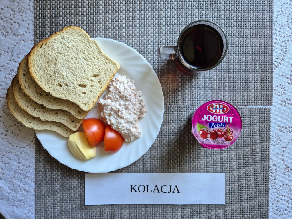 Na zdjęciu znajduję się posiłek w którego skład wchodzi Chleb pszenny, margaryna śniadaniowa, Pasta z sera białego i wędliny, Pomidor, jogurt owocowy oraz Herbata czarna
