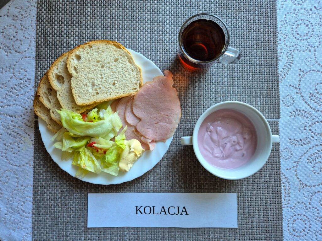 Na zdjęciu znajduję się posiłek w którego skład wchodzi Chleb pszenny, Margaryna Śniadaniowa, Wędlina wieprzowa, Sałatka z sałaty lodowej, pomidora i ogórka, koktajl owocowy oraz Herbata czarna