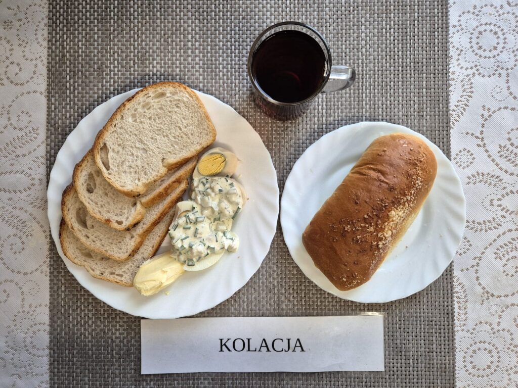 Na zdjęciu znajduję się posiłek w którego skład wchodzi Chleb pszenny, Margaryna Śniadaniowa, Jaja 2 szt w sosie tatarskim, Drożdzówka oraz Herbata czarna