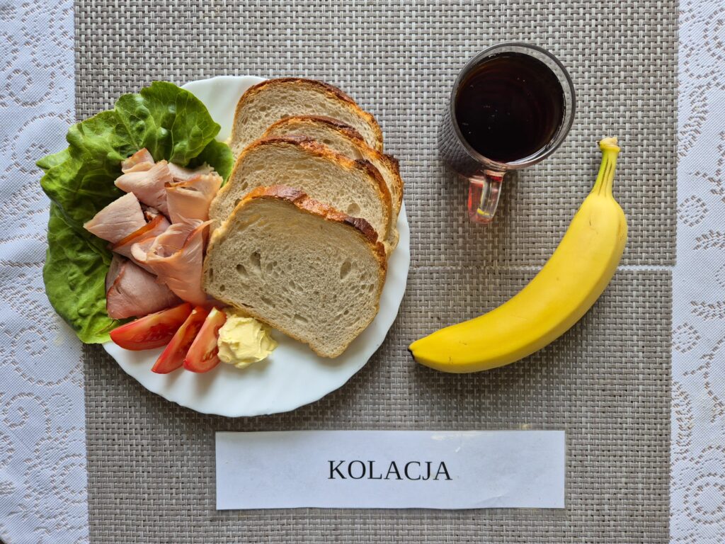 Na zdjęciu znajduję się posiłek w którego skład wchodzi Chleb pszenny, Margaryna Śniadaniowa, Wędlina wieprzowa, Sałata masłowa, Pomidor, banan oraz Herbata czarna