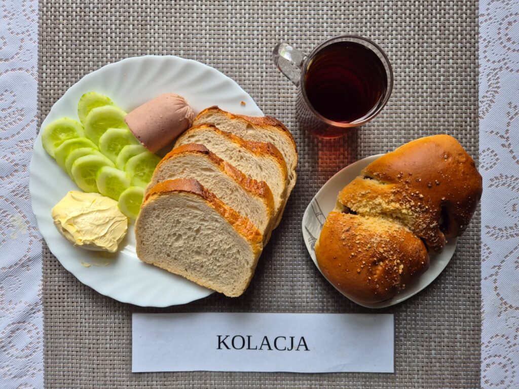 Na zdjęciu znajduję się posiłek w którego skład wchodzi Chleb pszenny, Margaryna Śniadaniowa, Pasztet drobiowy, pieczony, Ogórek zielony, Drożdżówka oraz Herbata czarna