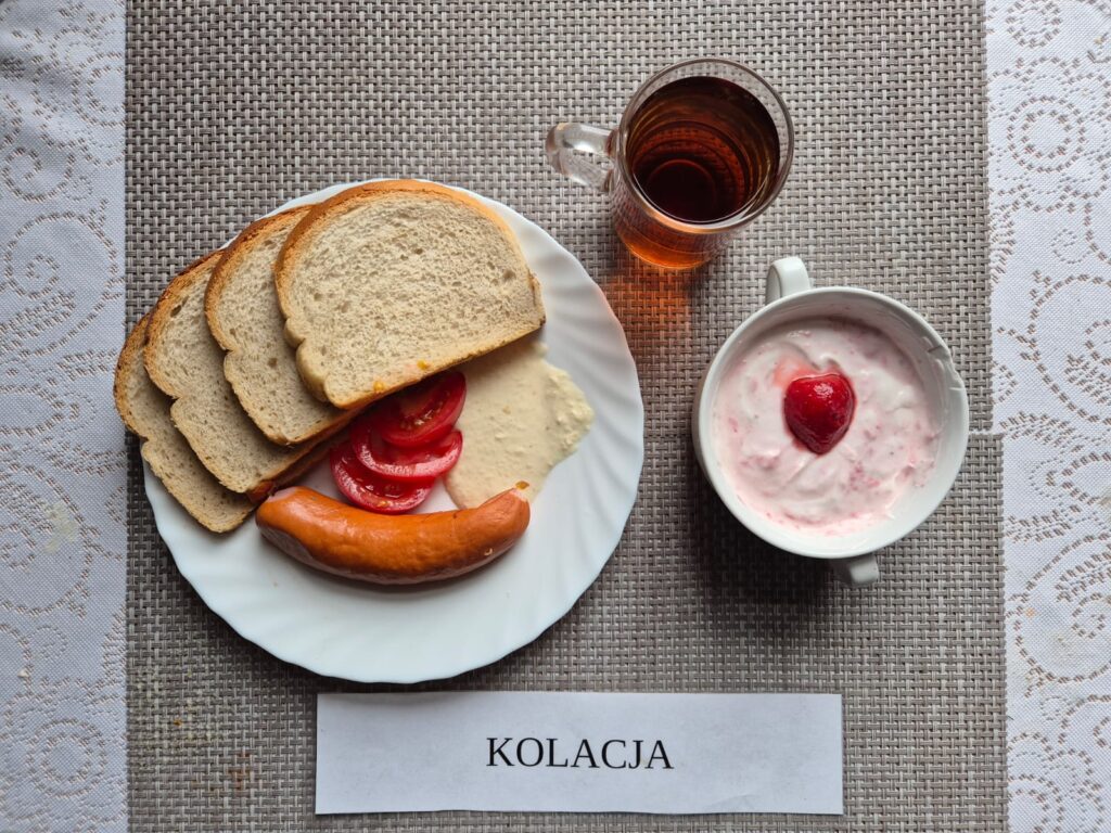 Na zdjęciu znajduję się posiłek w którego skład wchodzi Chleb pszenny, Margaryna Śniadaniowa, Kiełbasa parówkowa, Sos chrzanowy, koktajl owocowy oraz Herbata czarna