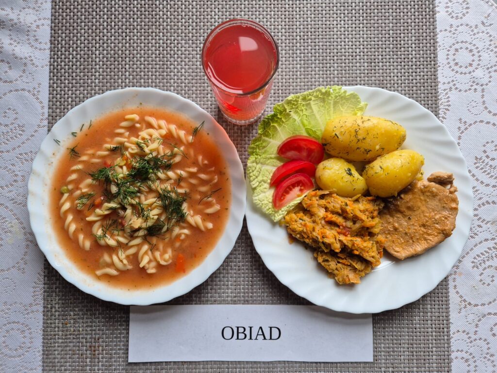 Na zdjęciu znajduję się posiłek w którego skład wchodzi Zupa pomidora z makaronem, Ziemniaki młode, Schab pieczony w sosie własnym, Kapusta młoda na ciepło oraz Kompot z jabłek