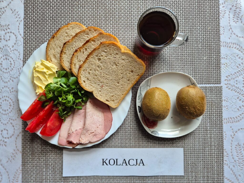 Na zdjęciu znajduję się posiłek w którego skład wchodzi Chleb pszenny, Margaryna Śniadaniowa, Szynka wieprzowa, Roszponka, pomidor oraz Herbata czarna