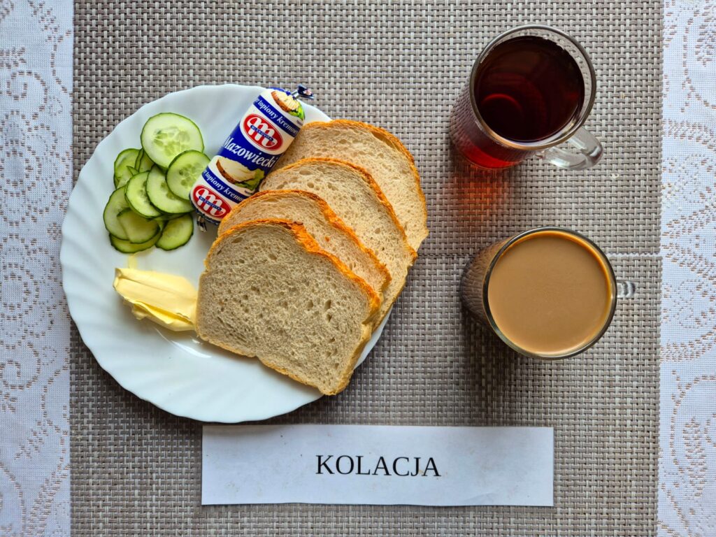 Na zdjęciu znajduję się posiłek w którego skład wchodzi Chleb pszenny, Margaryna Śniadaniowa, Ser topiony, Ogórek zielony, kakao oraz Herbata czarna