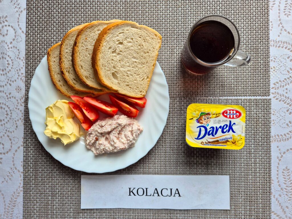 Na zdjęciu znajduję się posiłek w którego skład wchodzi Chleb pszenny, margaryna śniadaniowa, Pasta z twarogu i wędliny, pomidor, serek homogenizowany oraz Herbata czarna