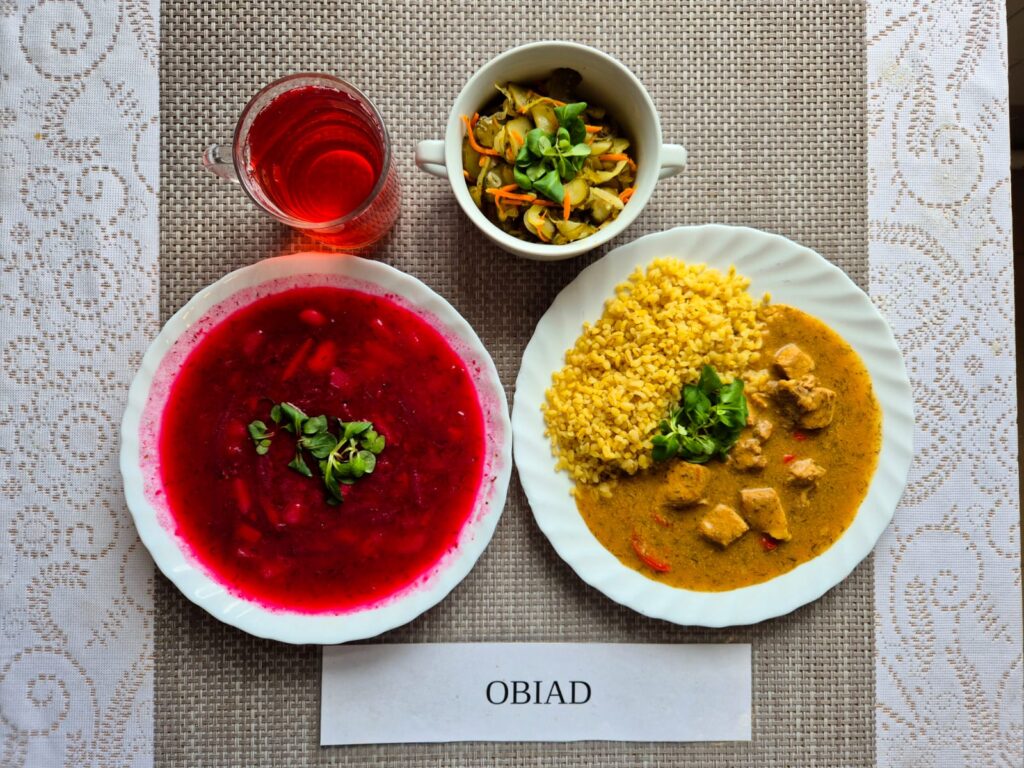 Na zdjęciu znajduję się posiłek w którego skład wchodzi Barszcz ukraiński z ziemniakami, Kasza bulgur, Gulasz wieprzowy, Surówka z ogórka kiszonego oraz Kompot z jabłek