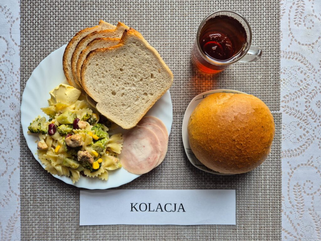 Na zdjęciu znajduję się posiłek w którego skład wchodzi Chleb pszenny, Margaryna Śniadaniowa, Sałatka makaronowa z suszonymi pomidorami i kurczakiem, bułka pszenna oraz Herbata czarna
