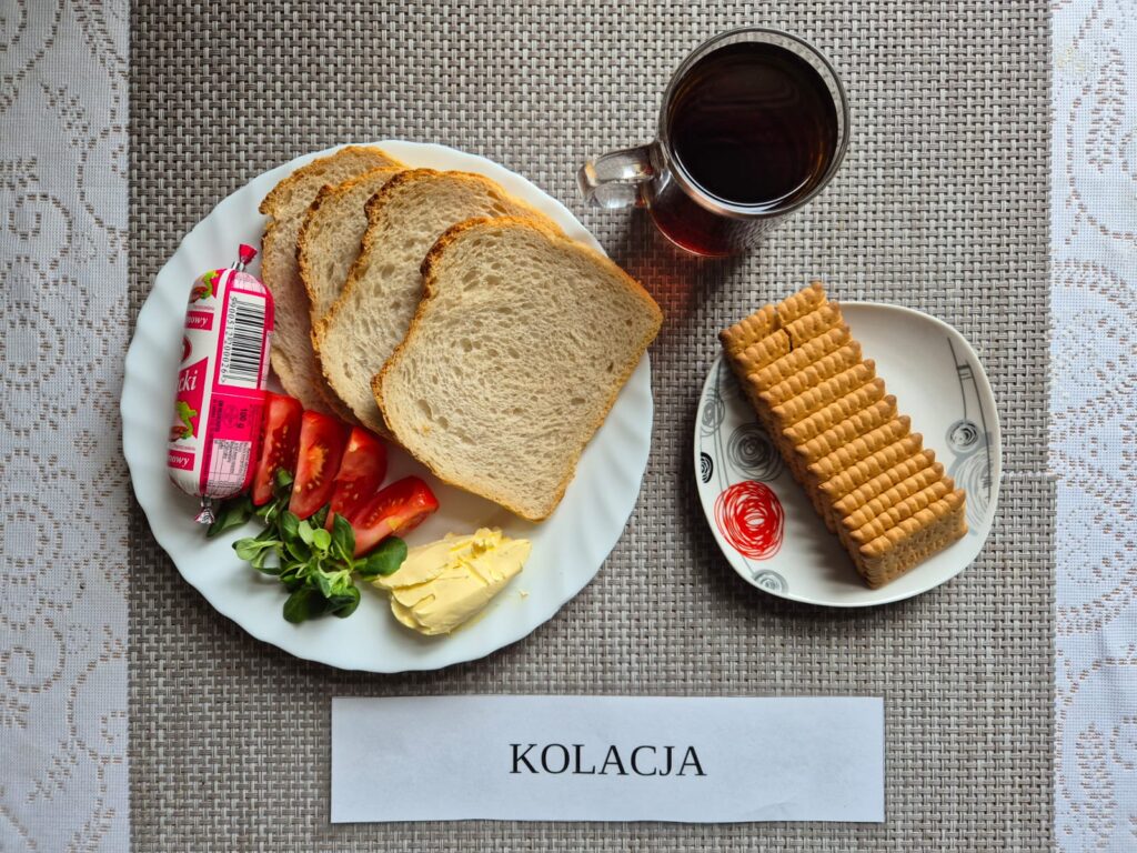 Na zdjęciu znajduję się posiłek w którego skład wchodzi Chleb pszenny, Margaryna Śniadaniowa, ser topiony, pomidor, herbatniki oraz Herbata czarna