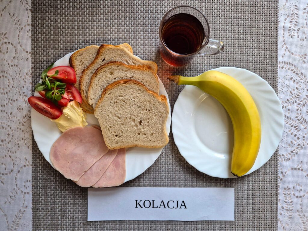 Na zdjęciu znajduję się posiłek w którego skład wchodzi Chleb pszenny, Margaryna Śniadaniowa, Kiełbasa krakowska wieprzowa, pomidor, banan oraz Herbata czarna