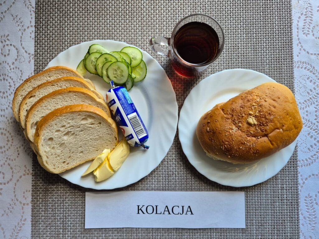Na zdjęciu znajduję się posiłek w którego skład wchodzi Chleb pszenny, Margaryna Śniadaniowa, Ser topiony, ogórek zielony, Drożdżówka oraz Herbata czarna