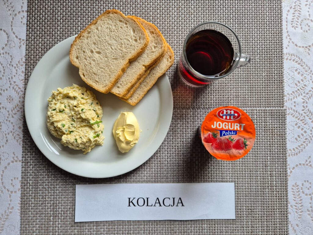 Na zdjęciu znajduję się posiłek w którego skład wchodzi Chleb pszenny, margaryna śniadaniowa, Pasta jajeczna ze szczypiorkiem, jogurt owocowy oraz Herbata czarna