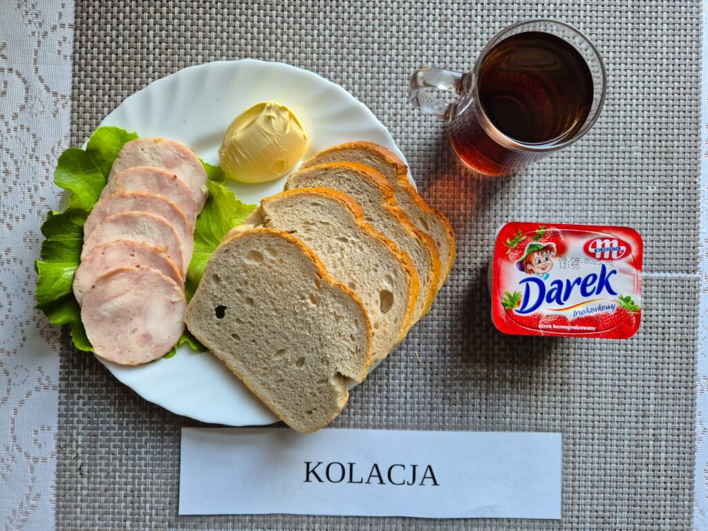 Na zdjęciu znajduję się posiłek w którego skład wchodzi Chleb pszenny, Margaryna Śniadaniowa, wędlina wieprzowa, serek homogenizowany oraz herbata czarna