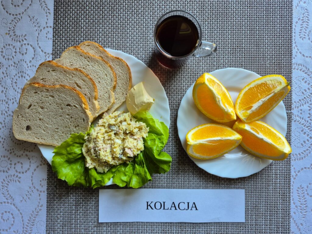 Na zdjęciu znajduję się posiłek w którego skład wchodzi Chleb pszenny, Margaryna Śniadaniowa, Pasta z tuńczyka, jajka i makaronu, sałata masłowa, pomarańcza oraz Herbata czarna